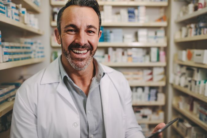 Smiling mature pharmacist with digital tablet standing by aisle in drug store