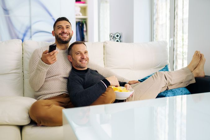 Male couple relaxing on sofa watching tv with bowl of chips