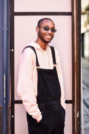Happy male in denim overalls and earrings with hands in pockets against a pink wall