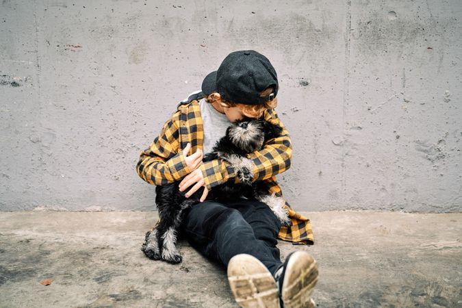 Child hugging dog while sitting near gray street wall