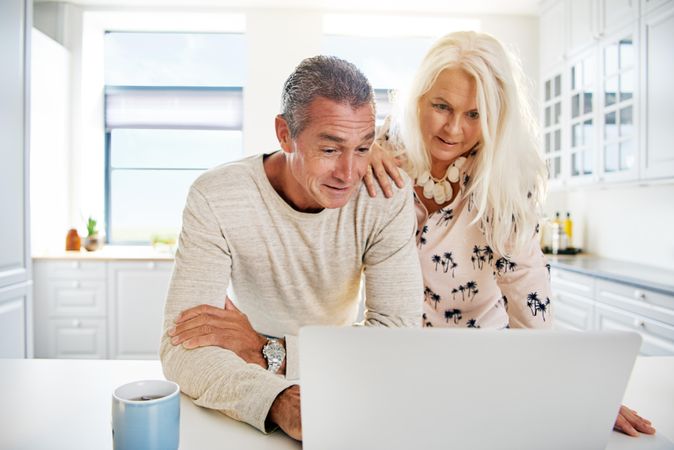 Mature woman looking at computer over man’s shoulder in kitchen