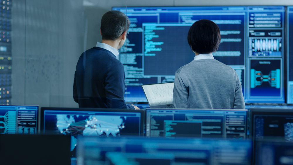In the System Control Room IT Specialist and Project Engineer Have Discussion, they're surrounded by Multiple Monitors with Graphics.