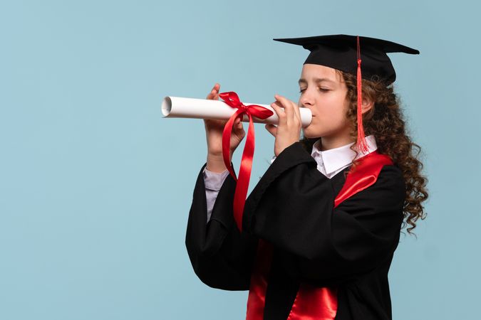 Girl in graduation attire pretending to play trumpet with diploma.