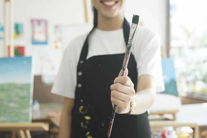 A close-up of the artist hands wearing an apron smeared with paint. Clutching many brushes and paintbrushes.