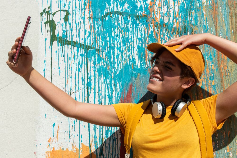 Trendy cool woman in yellow clothes taking selfie against painted wall in city