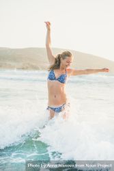 Woman in bikini jumping and smiling in the water at the beach with arms up and out 42nOy4