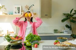 Smiling woman hiding her eyes with two lemons standing in the kitchen 5aMjG5