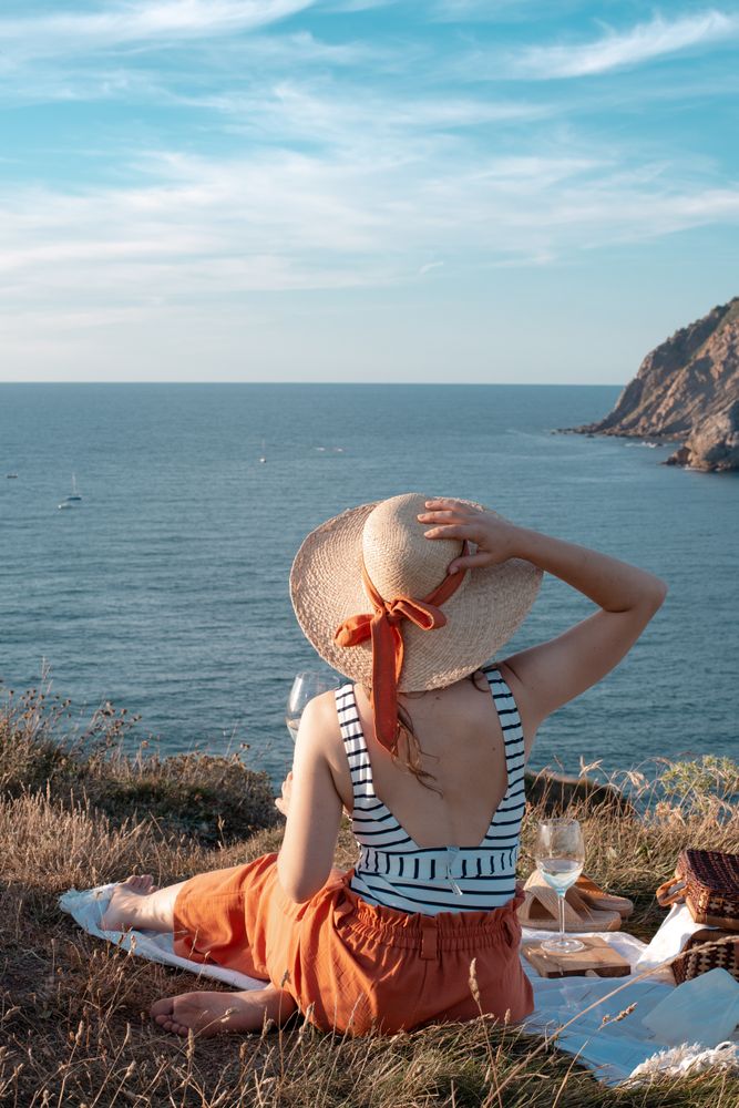 Woman with glass of drink on picnic mat looking at sea and mountains