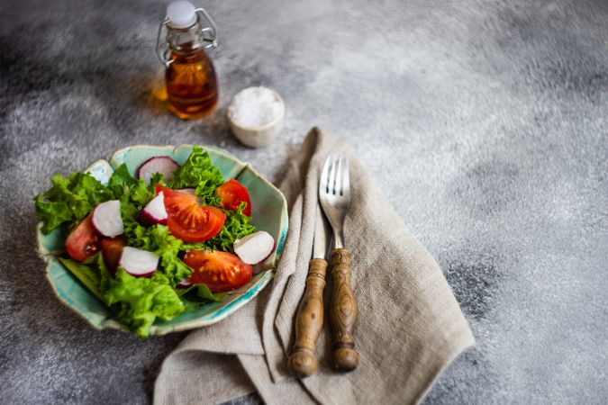 Bowl of salad with tomatoes, radish & lettuce on grey counter with oil, salt and copy space
