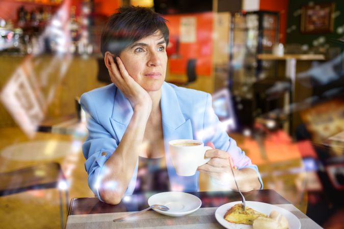 Female seen behind glass in trendy blue jacket sitting at table with cup of hot drink and slice of cake