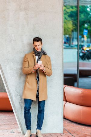 Coy man leaning on wall outside texting on phone and wearing a camel coat 