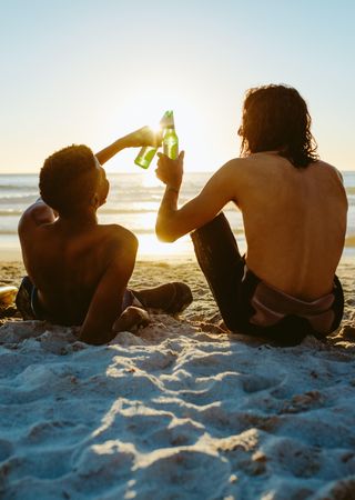 Rear view of two young friends sitting on beach and toasting beers at sunset