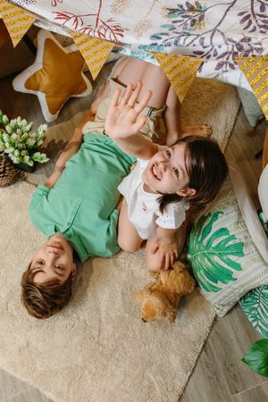 Happy little girl raising hand up and smiling boy looking while resting lying over rug in cozy tent.