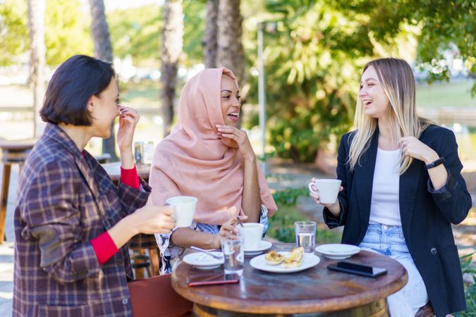 Three women laughing while enjoying coffee and baked goods on a beautiful sunny day
