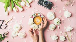 Coffee, camera, tulips artfully arranged on pink background, with hands on coffee cup