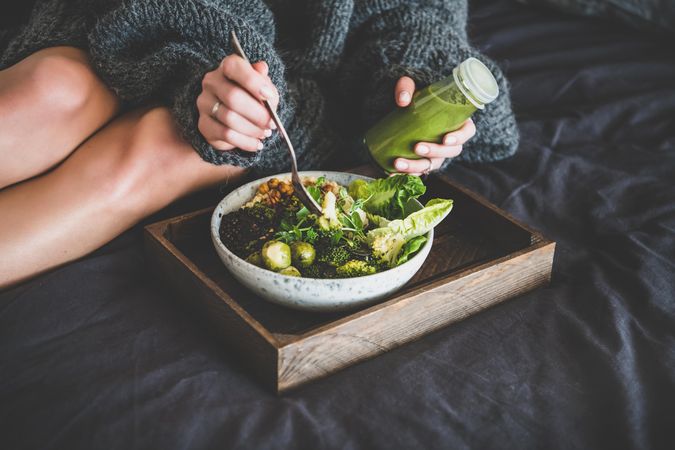 Woman in cozy sweater sitting with wooden tray of vegetarian bowl with smoothie on side