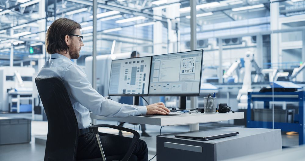 Software Developer Programming an Artificial Intelligence Neural Network on a Desktop Computer. Male Working in a Modern Technological Facility with an Automated Production Line with Robotic Arms