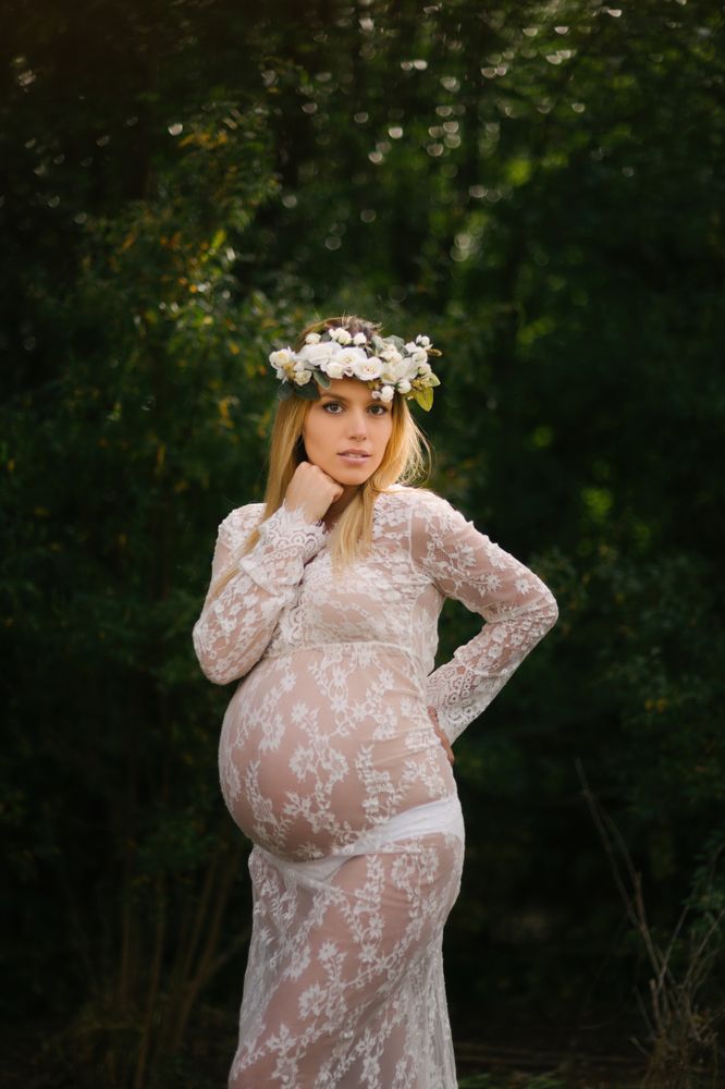 Pregnant woman with closed eyes in white lace dress touching chin while standing in forest