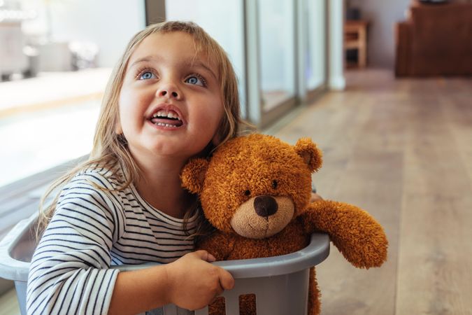 Adorable young girl sitting in a laundry basket with her teddy bear