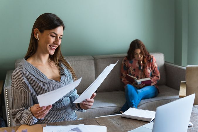 Cheerful woman reading documents and sitting at desk with laptop