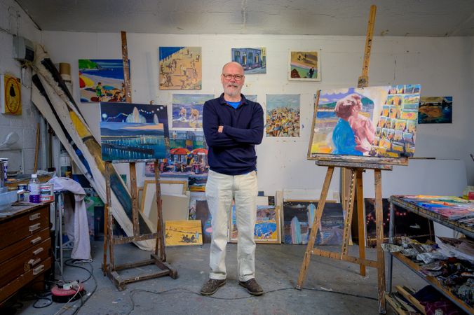 Happy older male with arms crossed surrounded by paintings in art studio