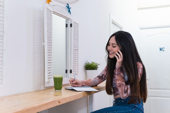 Woman taking notes in notebook and speaking on mobile phone sitting in bar with healthy beverage