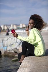 Smiling female with curly hair in bright green shirt sitting listening to music on dock