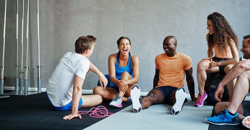 Group of people in workout gear sitting on gym floor sharing a laugh