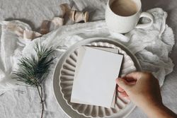 Hand placing blank card, invitation mockup on ceramic plate with pine branch on tablecloth