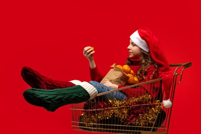 Girl in Santa hat sitting in shopping cart holding oranges.