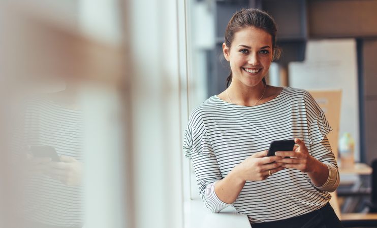 Smiling businesswoman holding mobile phone standing in office