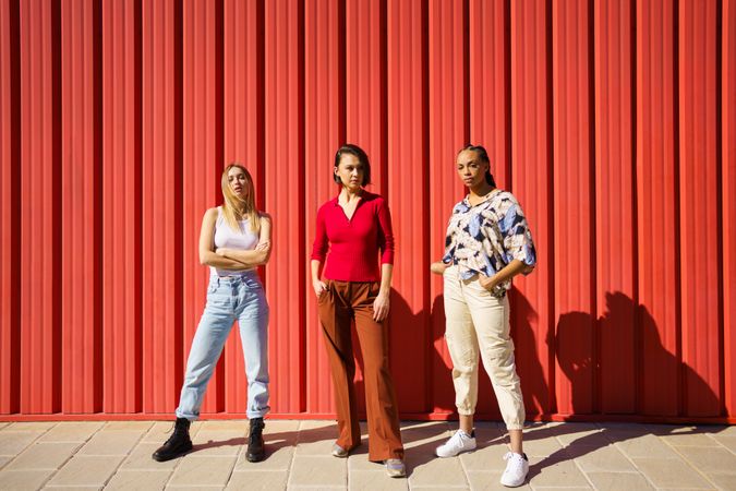 Three serious women posing in front of red wall on sunny day
