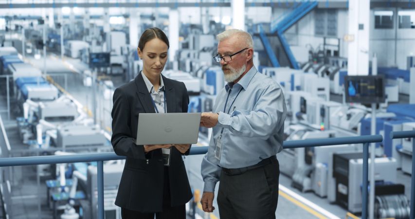 Experienced Male and Female Engineers Standing on a Platform at a Modern Industrial Factory, Using Laptop Computer and Discussing Production of Modern Electronics for Artificial Intelligence Machines