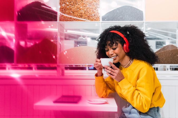 Cheerful Black woman in headphones drinking coffee in modern cafeteria