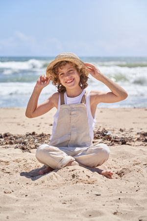Smiling boy sitting on sandy beach and touching straw hat