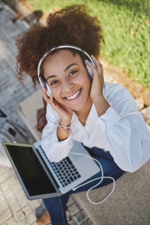 Cheerful Black woman listening to music and using laptop on stre