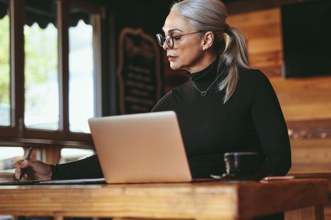 Mature female at cafe with laptop writing on notepad