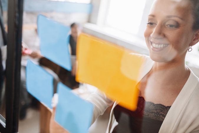 Smiling young business woman sticking adhesive notes on glass wall