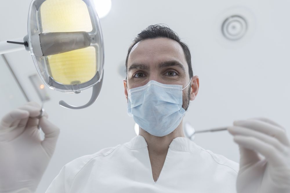 Dentist wearing surgical mask while holding angled mirror and drill, ready to begin