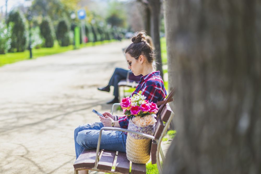 Side view of a young hipster woman sitting on a park bench relaxing in a sunny day while using a mobile phone