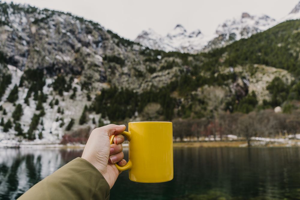 Person with cup near wonderful lake between hills in snow and cloudy sky