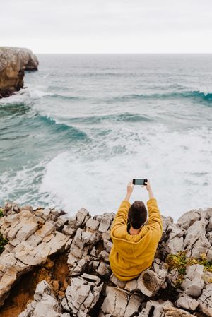 Woman in yellow hoodie taking selfie on smartphone on rocky shore