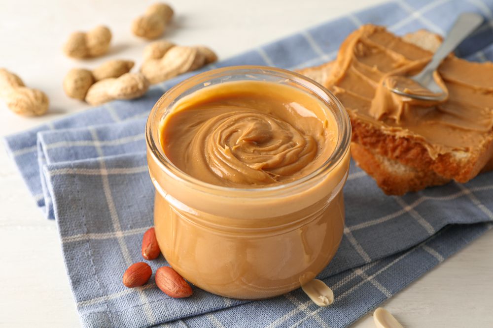 Glass jar with peanut butter, peanut, kitchen towel, spoon and peanut butter sandwich on wooden background, space for text and closeup