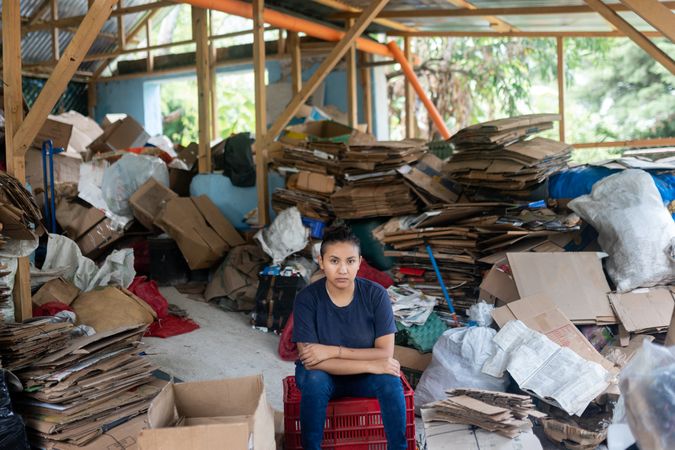 Young Hispanic Woman Determined Among Cardboard for Recycling in Warehouse. Serious Portrait