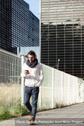 Young man walking in scarf past metallic fence and holding smartphone outdoors 0yXPAa