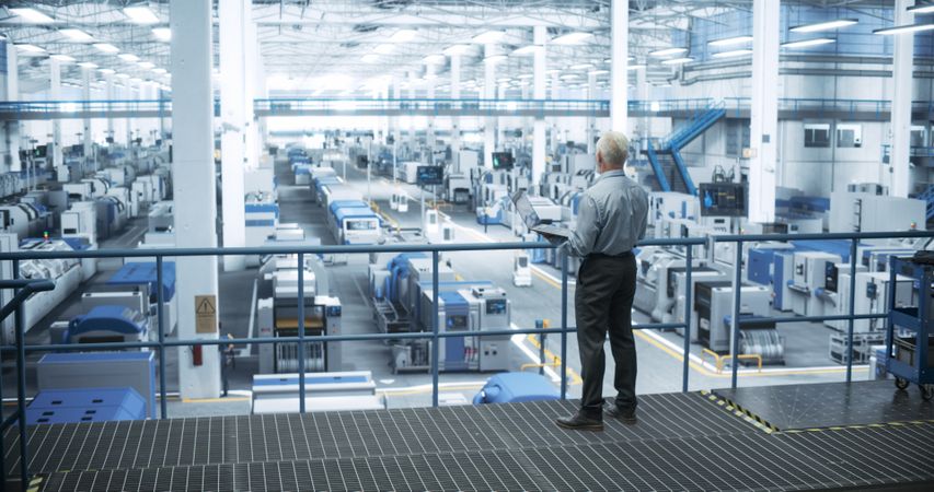 Factory Manager Working on Laptop at a Futuristic Industrial Manufacture with Autonomous AI Robots. Specialist Monitoring All the Data on a Computer, Checking Reports and Planning Workload