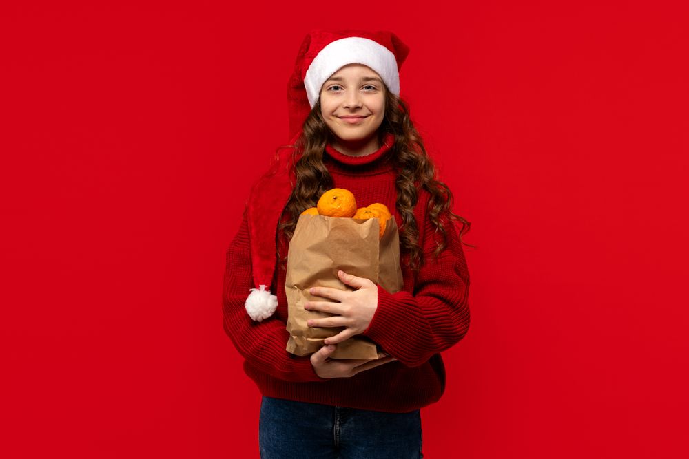 Girl in Santa hat holds paper bag filled with oranges.
