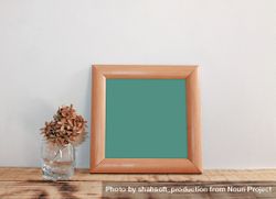 Plain square wooden picture frame with green interior leaning against wall mockup with cup of dried flowers 4ONvJ5