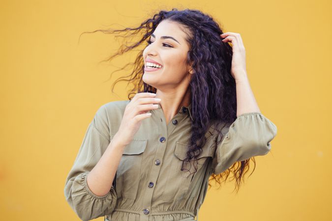 Female in army green jumpsuit smiling in front of mustard wall touching with hair back