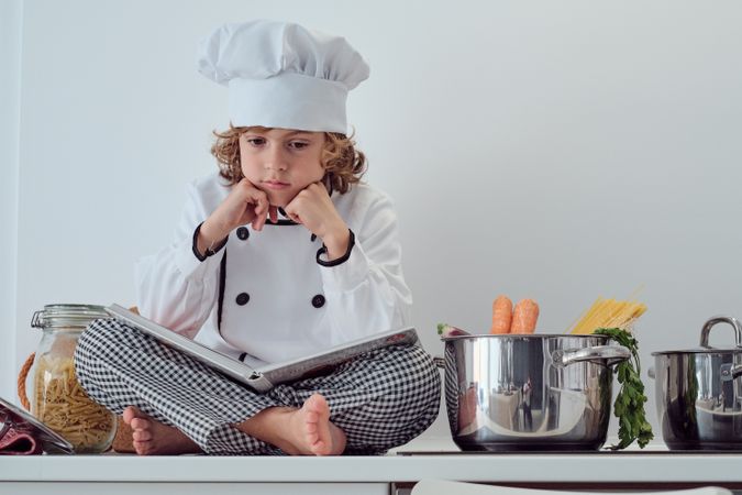 Pensive preteen boy in chef uniform sitting with opened cookbook